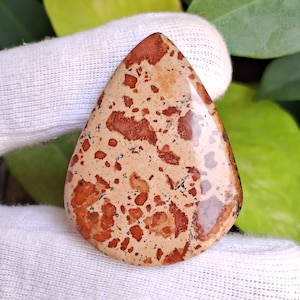 May include: A teardrop-shaped gemstone with a beige base and scattered reddish-brown spots. The polished surface reflects light, highlighting the natural patterns. The stone is likely used for jewelry or decorative purposes.