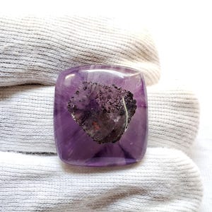 Puede incluir: Una piedra preciosa de amatista cuadrada y pulida con una inclusión oscura e irregular en el centro. La piedra preciosa es de color púrpura intenso con vetas más claras y un acabado brillante. La inclusión es una mezcla de gris oscuro y negro.
