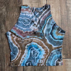 Pode incluir: Um top curto sem mangas com um padr&atilde;o tie-dye de geodo em tons de azul, castanho e branco. A pe&ccedil;a tem um decote alto e uma bainha reta, com faixas de cor circulares conc&ecirc;ntricas que imitam a apar&ecirc;ncia de forma&ccedil;&otilde;es rochosas naturais.