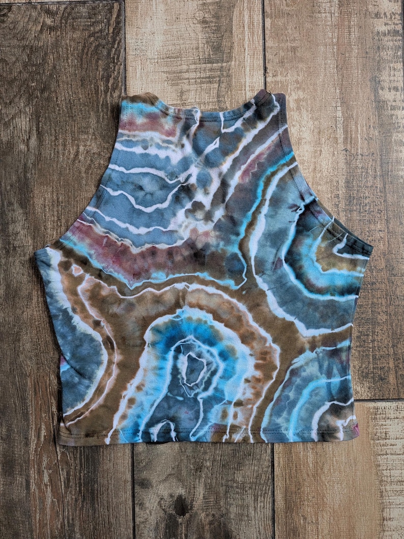 Pode incluir: Um top curto sem mangas com um padr&atilde;o tie-dye de geodo em tons de azul, castanho e branco. A pe&ccedil;a tem um decote alto e uma bainha reta, com faixas de cor circulares conc&ecirc;ntricas que imitam a apar&ecirc;ncia de forma&ccedil;&otilde;es rochosas naturais.