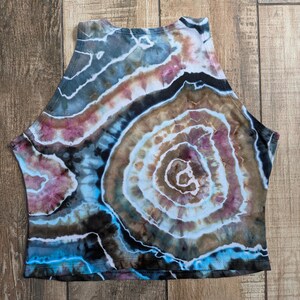 Pode incluir: Um top curto sem mangas com um padr&atilde;o de geodo tie-dye em tons de castanho, preto, azul e rosa suave. O design consiste em an&eacute;is circulares conc&ecirc;ntricos que irradiam para fora, criando uma apar&ecirc;ncia de pedra no tecido.