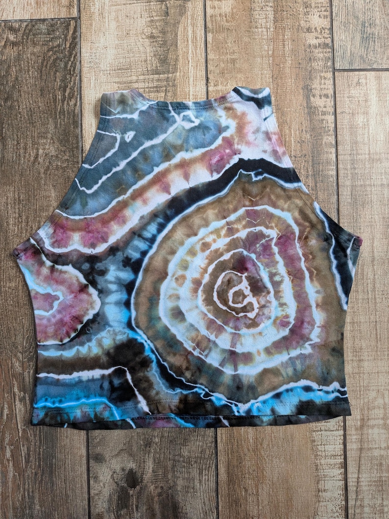Pode incluir: Um top curto sem mangas com um padr&atilde;o de geodo tie-dye em tons de castanho, preto, azul e rosa suave. O design consiste em an&eacute;is circulares conc&ecirc;ntricos que irradiam para fora, criando uma apar&ecirc;ncia de pedra no tecido.