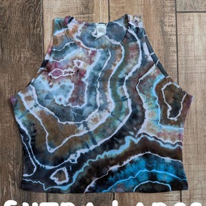 Pode incluir: Um top curto sem mangas com um padr&atilde;o tie-dye em tons de azul, castanho, preto e branco, que lembra uma geode. A pe&ccedil;a tem um decote alto e uma etiqueta na gola. O texto EXTRA LARGE est&aacute; impresso a branco na parte inferior.