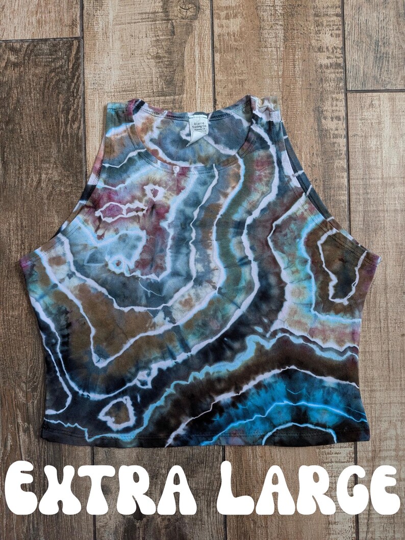 Pode incluir: Um top curto sem mangas com um padr&atilde;o tie-dye em tons de azul, castanho, preto e branco, que lembra uma geode. A pe&ccedil;a tem um decote alto e uma etiqueta na gola. O texto EXTRA LARGE est&aacute; impresso a branco na parte inferior.