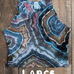 Pode incluir: Um top curto sem mangas com um padr&atilde;o de geodo tie-dye em tons de azul, castanho, preto e branco. A pe&ccedil;a tem um decote alto e uma etiqueta interior branca na gola. A palavra LARGE est&aacute; impressa a branco na parte inferior da imagem.