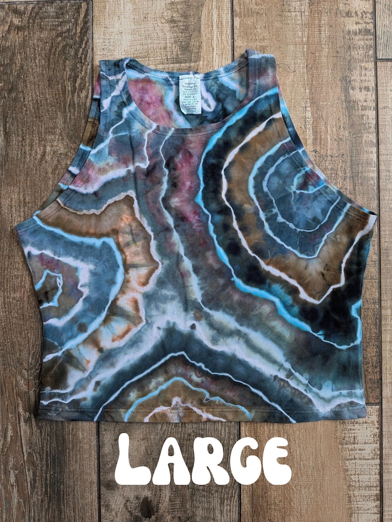 Pode incluir: Um top curto sem mangas com um padr&atilde;o de geodo tie-dye em tons de azul, castanho, preto e branco. A pe&ccedil;a tem um decote alto e uma etiqueta interior branca na gola. A palavra LARGE est&aacute; impressa a branco na parte inferior da imagem.