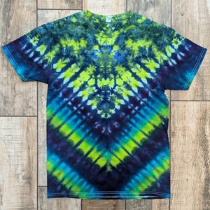 Pode incluir: Uma camiseta de manga curta com um padrão tie-dye em tons de azul, verde e amarelo. O design apresenta um padrão de chevron que irradia do centro da camiseta. A camiseta é feita de um material macio.
