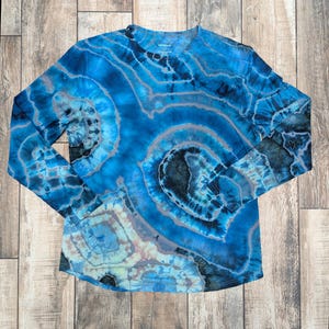 Pode incluir: Uma camisa de manga comprida com gola redonda e um padrão tie-dye vibrante em tons de azul, cinza e preto. O design da camisa apresenta círculos concêntricos e padrões em espiral, criando um visual único e artístico. A camisa é feita de um material macio e confortável.