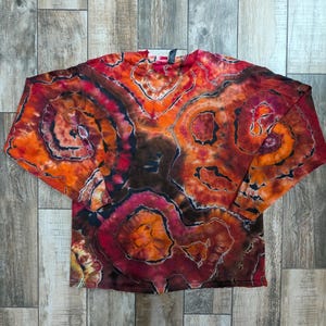 Pode incluir: Uma camisa tie-dye de manga comprida em tons de laranja, vermelho e castanho. A camisa apresenta um design de padrão em espiral. A peça de roupa está estendida numa superfície de madeira.
