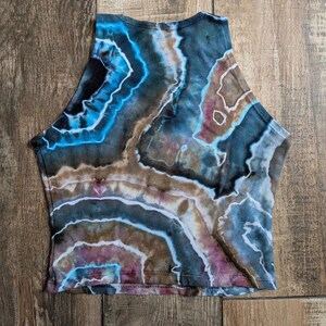 Pode incluir: Um top curto sem mangas com um padr&atilde;o tie-dye inspirado em geodos em tons de azul profundo, castanho, preto e toques de roxo. O design consiste em formas org&acirc;nicas irregulares e conc&ecirc;ntricas com linhas brancas a separar as cores.