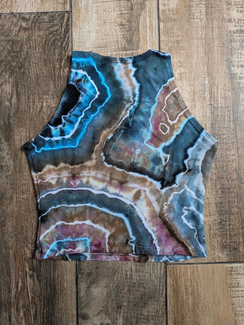 Pode incluir: Um top curto sem mangas com um padr&atilde;o tie-dye inspirado em geodos em tons de azul profundo, castanho, preto e toques de roxo. O design consiste em formas org&acirc;nicas irregulares e conc&ecirc;ntricas com linhas brancas a separar as cores.