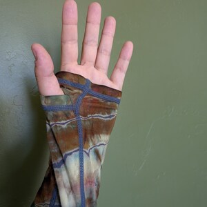 May include: A long-sleeved garment featuring a tie-dye pattern in shades of brown, cream, and muted blue. The sleeve includes a thumb hole opening at the cuff, finished with visible dark blue flatlock stitching along the seams and edges.