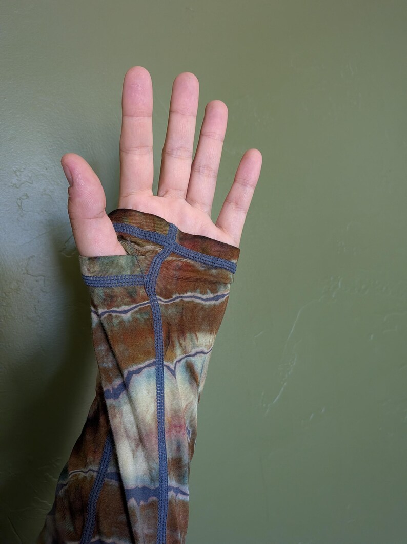 May include: A long-sleeved garment featuring a tie-dye pattern in shades of brown, cream, and muted blue. The sleeve includes a thumb hole opening at the cuff, finished with visible dark blue flatlock stitching along the seams and edges.