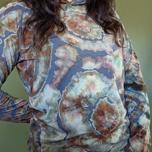 May include: A long-sleeve hooded shirt featuring a tie-dye pattern in shades of slate blue, earthy brown, and muted teal. The garment has a hood, long sleeves with thumb holes, and a circular, organic-looking dye design across the fabric.