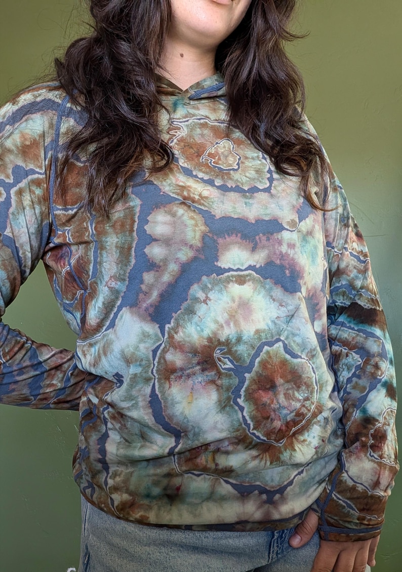 May include: A long-sleeve hooded shirt featuring a tie-dye pattern in shades of slate blue, earthy brown, and muted teal. The garment has a hood, long sleeves with thumb holes, and a circular, organic-looking dye design across the fabric.