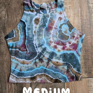 Pode incluir: Um top curto sem mangas com um padr&atilde;o tie-dye de faixas rodopiantes em azul, castanho, branco e magenta. A pe&ccedil;a tem um decote alto e uma etiqueta interior branca na gola. A palavra MEDIUM est&aacute; impressa a branco na parte inferior.
