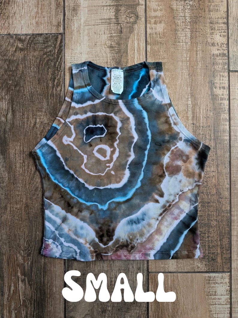 Pode incluir: Um top curto sem mangas com um padr&atilde;o tie-dye inspirado em geodos em tons de castanho, azul e branco. A pe&ccedil;a tem um decote alto e uma etiqueta interior branca na gola. A palavra SMALL est&aacute; impressa a branco na parte inferior.