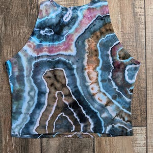 Pode incluir: Um top curto sem mangas com um padr&atilde;o tie-dye de faixas rodopiantes em azul, castanho, rosa e branco. A pe&ccedil;a tem um decote alto e uma bainha reta, exibindo uma est&eacute;tica semelhante a uma geoda com an&eacute;is de cor conc&ecirc;ntricos no tecido.