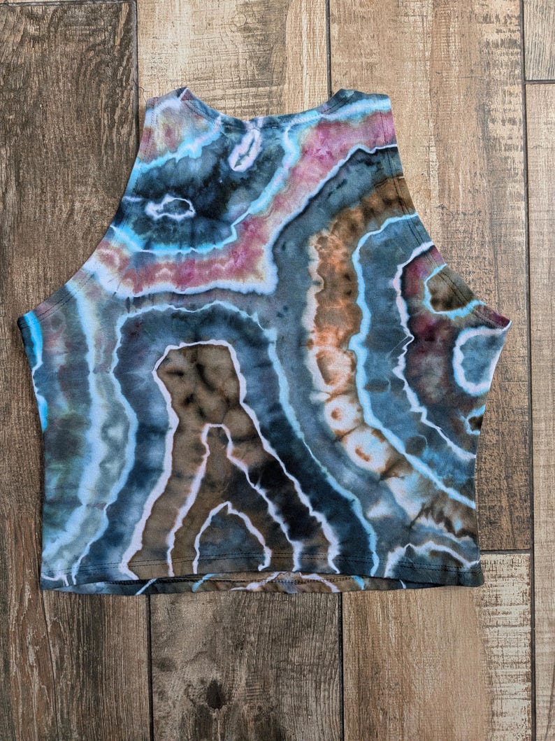 Pode incluir: Um top curto sem mangas com um padr&atilde;o tie-dye de faixas rodopiantes em azul, castanho, rosa e branco. A pe&ccedil;a tem um decote alto e uma bainha reta, exibindo uma est&eacute;tica semelhante a uma geoda com an&eacute;is de cor conc&ecirc;ntricos no tecido.