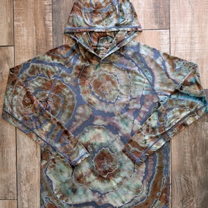May include: A long-sleeve hooded shirt featuring a tie-dye pattern in shades of brown, sage green, and muted blue. The garment has a hood, raglan sleeves, and visible seam detailing throughout, creating a circular, organic, and layered aesthetic.