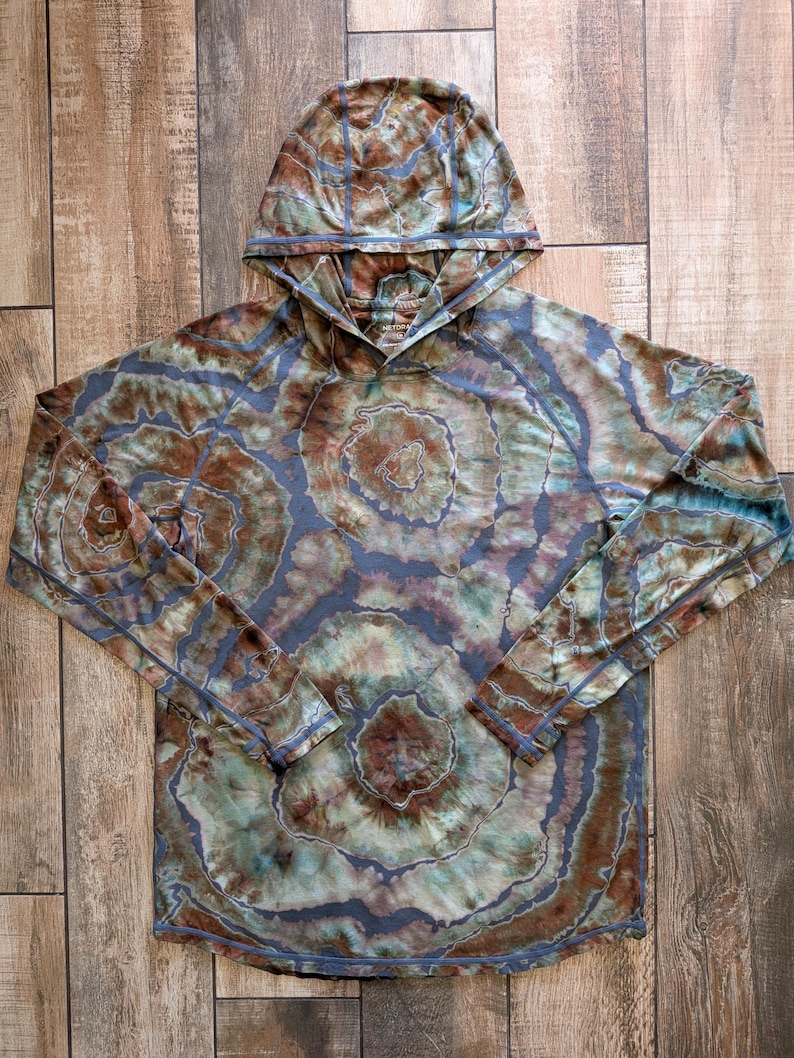 May include: A long-sleeve hooded shirt featuring a tie-dye pattern in shades of brown, sage green, and muted blue. The garment has a hood, raglan sleeves, and visible seam detailing throughout, creating a circular, organic, and layered aesthetic.