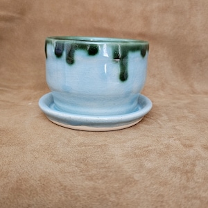 May include: A light blue ceramic planter with a green glaze dripping down the sides. The planter has a matching saucer.