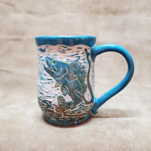 May include: A blue ceramic mug with a white glaze and a raised design of a fish jumping out of the water. The mug has a handle and is perfect for enjoying a hot beverage.