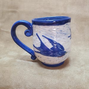 May include: A blue and white ceramic mug with a blue bird design. The mug has a handle and a rounded shape.