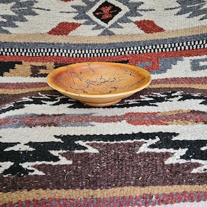 May include: A wooden bowl with a natural wood grain pattern and black lines. The bowl is sitting on a colorful rug with a geometric design.