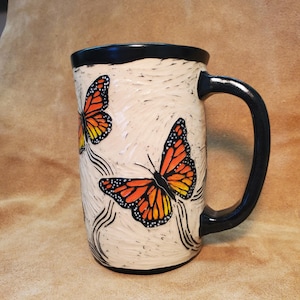 May include: A ceramic mug with a textured off-white surface featuring two painted monarch butterflies with orange, black, and white wings. The mug has a solid black rim and a matching black handle, with carved black lines accenting the butterflies.