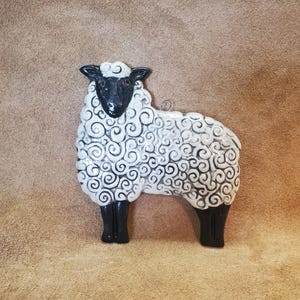 May include: A ceramic sheep figurine with a black head, legs, and a white body with a swirling pattern. The sheep is standing upright and has a loop for hanging. The background is a solid brown color.