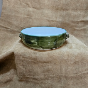 May include: A green ceramic bowl with two small handles on the sides. The bowl has a white interior and a green glaze with a mottled pattern.