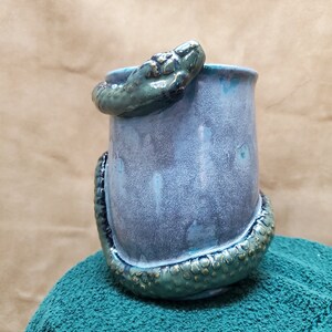 May include: A ceramic mug with a green snake wrapped around it. The mug is glazed in a light blue color with a white speckled finish.