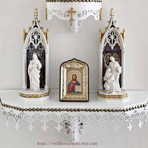 May include: A white wall-mounted religious display featuring a crucifix, two gothic-style niches with statues, and a framed icon. The display has ornate detailing with gold accents and is mounted on a white wall.