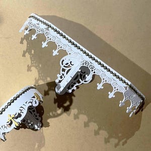 May include: Two white decorative wall shelves with intricate, openwork designs. The shelves have a crown-like edge with a dark trim and gold accents. The shelves are displayed on a neutral background, casting shadows.