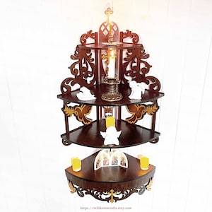 May include: A dark brown, three-tiered corner shelf with ornate carvings and gold accents. The shelf holds candles, small angel figurines, and an icon. A lit candle sits on the top shelf, with a cross at the top.