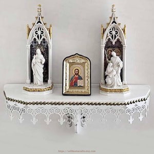 May include: A white wall-mounted shelf with ornate scalloped trim and gold bead accents. Two white and gold gothic-style arched shrines house religious figurines, flanking a central rectangular icon featuring a painted figure of Jesus Christ.