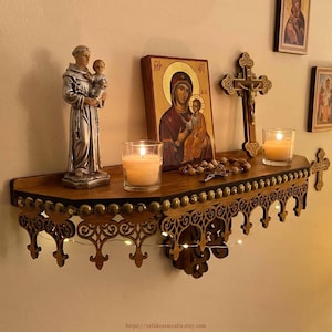 May include: A wooden shelf displaying religious items: a statue, an icon, a crucifix, and candles. The shelf has a decorative, dark brown, carved edge and is decorated with small lights. The items are arranged against a light-coloured wall.