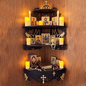 Op de afbeelding: Een zwart houten hoekplank met drie niveaus en gouden accenten, met religieuze iconen, kaarsen, een boek en rozenkransen. De plank heeft een kruis aan de boven- en onderkant, met decoratieve gravures.