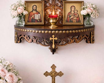May include: A dark wood wall shelf with ornate carvings, holding two framed religious icons, a lit candle, and bouquets of pink roses. A gold cross hangs below the shelf, with a decorative cross on a stand in the foreground. The shelf is decorated with gold accents.