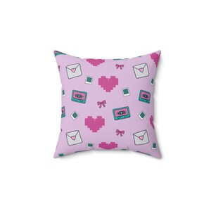 May include: Square pillow with a repeating pattern of pixelated hearts, cassette tapes, envelopes with hearts, bows, and small square and diamond shapes. The background is a light purple color, and the design elements are in shades of pink, teal, and white.