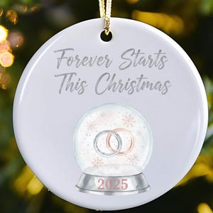 May include: A white ceramic Christmas ornament with the text "Forever Starts This Christmas." It features a snow globe design with two rings and the year 2025. A gold string is attached for hanging.