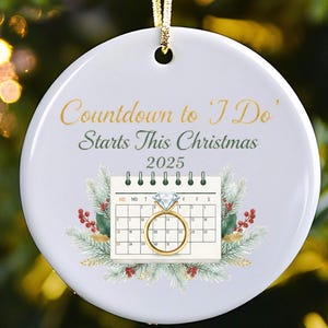 May include: White ceramic Christmas ornament with gold text reading "Countdown to 'I Do' Starts This Christmas 2025." Features a calendar with a diamond ring and festive greenery. Includes a gold hanging cord.