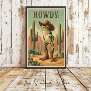 Könnte beinhalten: Ein gerahmter Kunstdruck mit einem grünen Frosch, der einen Cowboyhut und Stiefel trägt, mit dem Wort "HOWDY." darüber. Der Hintergrund zeigt Kakteen und eine Wüstenlandschaft. Der Druck ist vor einer rustikalen Holzwand platziert.