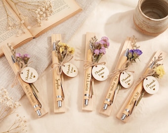 Puede incluir: Cinco bolígrafos de madera clara, cada uno montado sobre una base de madera a juego. Cada bolígrafo está decorado con un pequeño ramo de flores secas atado con cuerda y una etiqueta redonda de madera grabada con el texto Thank You y un corazón.