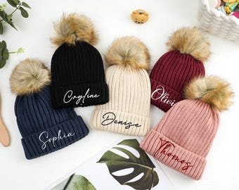 May include: Five knit beanies with faux fur pom poms. Each beanie has a different name embroidered on the front in white or red thread. The names are:  Olivia, Denise, Thomas, Sophia, and  Crayline.