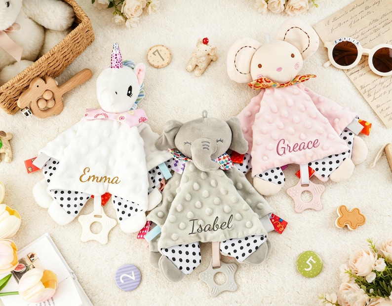 Puede incluir: Tres mantitas de apego de peluche con forma de unicornio blanco, elefante gris y rat&oacute;n rosa. Cada manta tiene una textura de puntos, una parte inferior con lunares blancos y negros, un mordedor en forma de estrella y los nombres bordados Emma, Isabel y Greace.