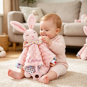 Puede incluir: Una manta de seguridad de peluche rosa con forma de conejo, con tela texturizada de puntos, una pajarita de lunares coloridos y un mordedor en forma de estrella en la parte inferior. El nombre Emma est&aacute; bordado en cursiva en la manta.