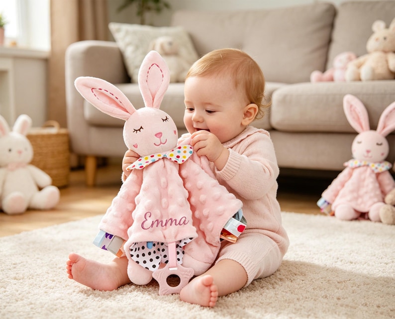 Puede incluir: Una manta de seguridad de peluche rosa con forma de conejo, con tela texturizada de puntos, una pajarita de lunares coloridos y un mordedor en forma de estrella en la parte inferior. El nombre Emma est&aacute; bordado en cursiva en la manta.
