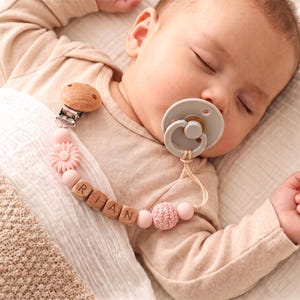 May include: A pacifier clip featuring a round wooden clip, light pink silicone beads, a flower-shaped bead, a crocheted bead, and wooden letter blocks spelling the name RIAN, attached to a light gray pacifier with a circular handle.